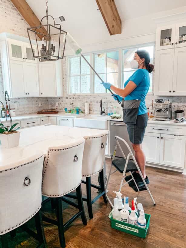 professional maid cleaning a hanging lamp above a kitchen table